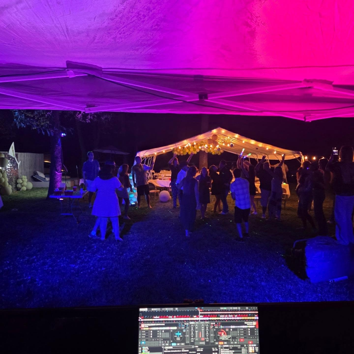 View from the DJ booth at a backyard birthday — tent, pink and purple lighting, guests dancing with glow sticks