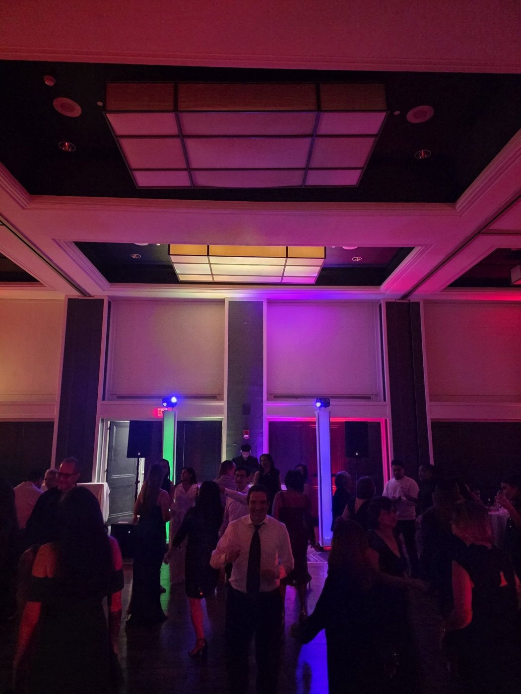 Packed ballroom dance floor with green, blue and pink uplighting totems and a glowing coffered ceiling