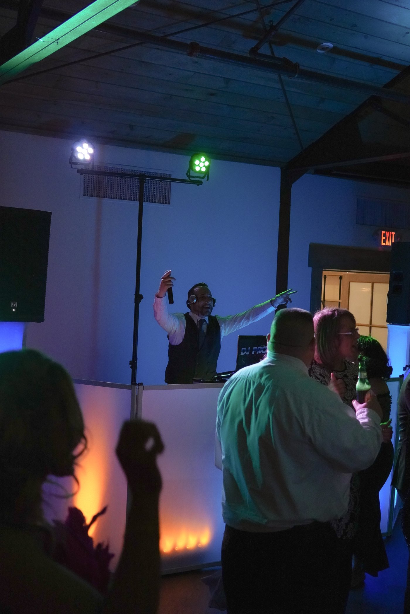 Barry with microphone raised at the booth, green and purple lights, guests dancing
