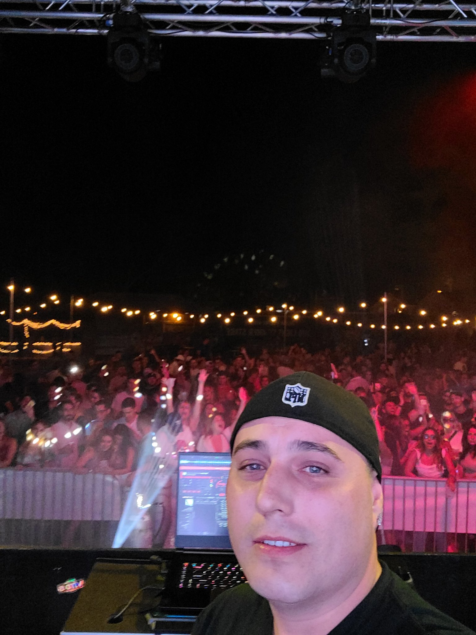 Barry taking a selfie from the DJ booth at an outdoor night event — huge crowd with hands up under string lights