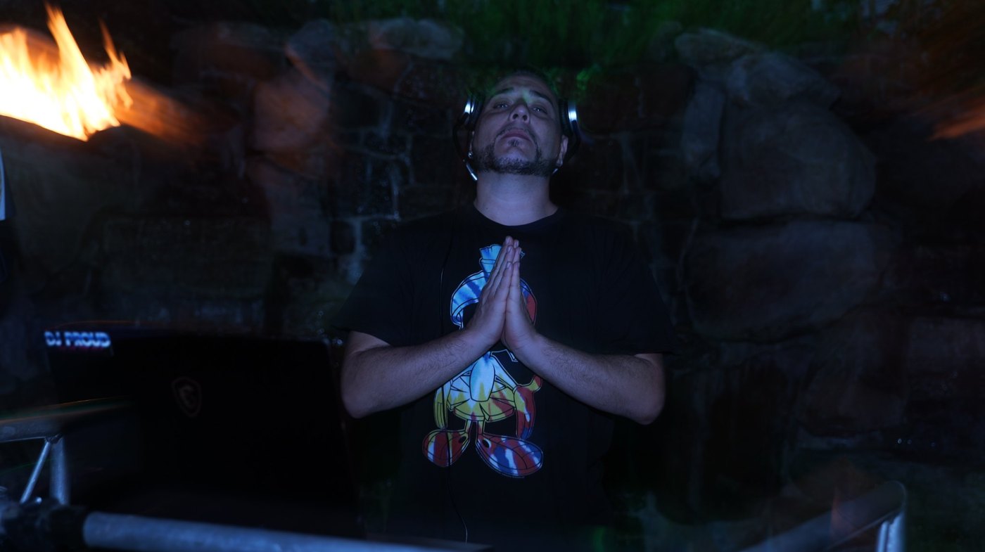 Barry with hands together and headphones on at an outdoor night gig, flame feature glowing beside the booth