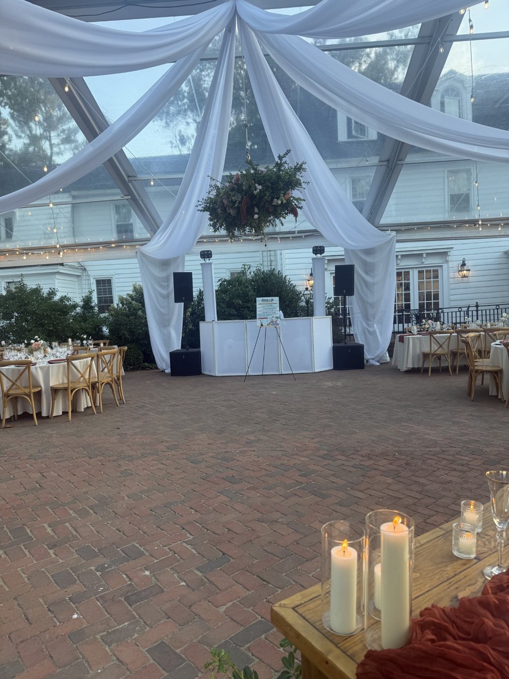 White DJ facade with speakers and uplighting set up under a draped clear tent at an outdoor wedding