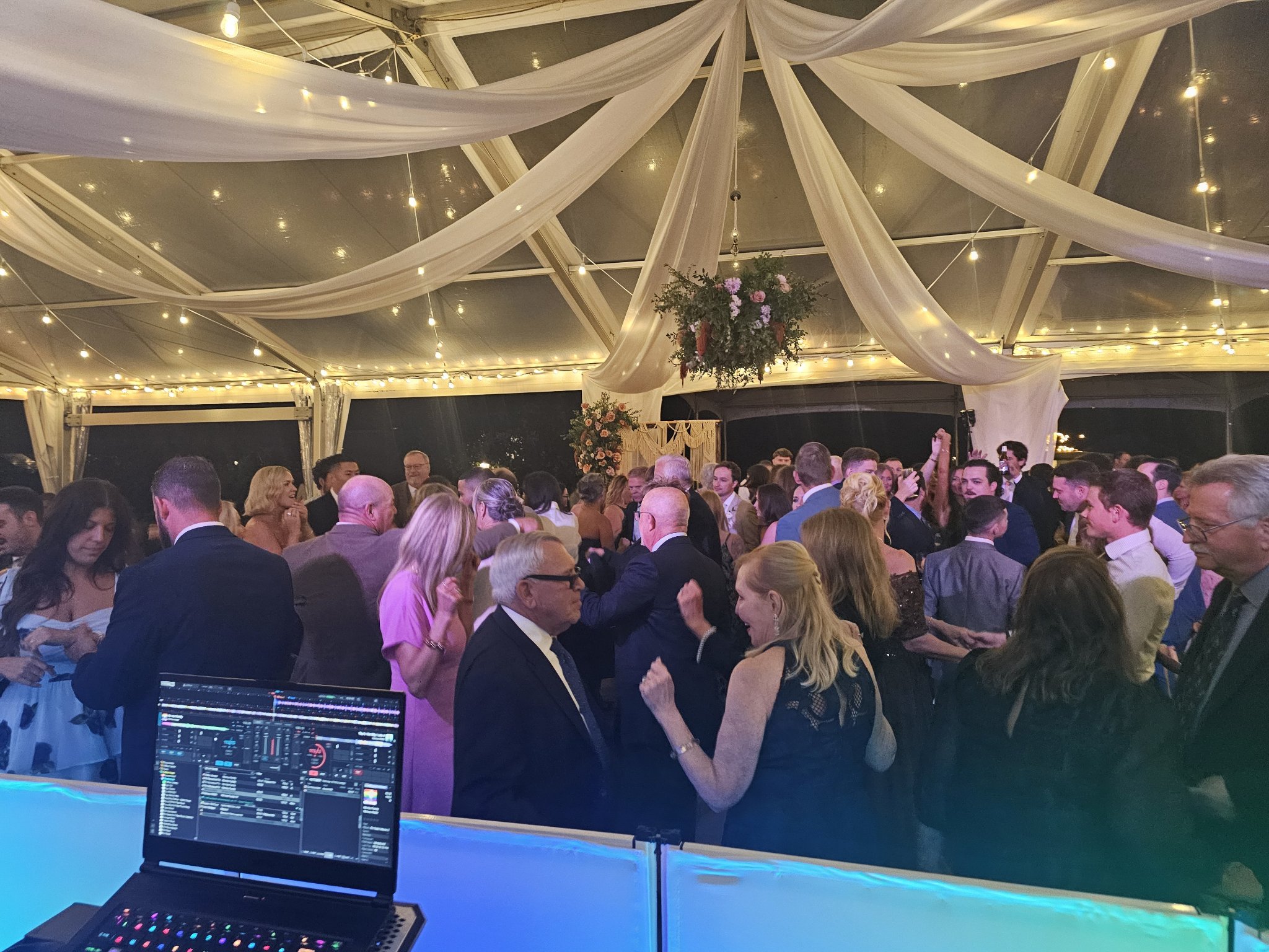 View from behind the DJ booth — laptop with DJ software and a packed wedding dance floor under the tent