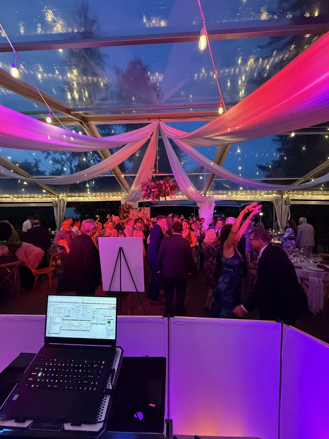 View from behind the DJ booth — laptop and glowing facade with a packed dance floor under a clear tent lit pink and purple