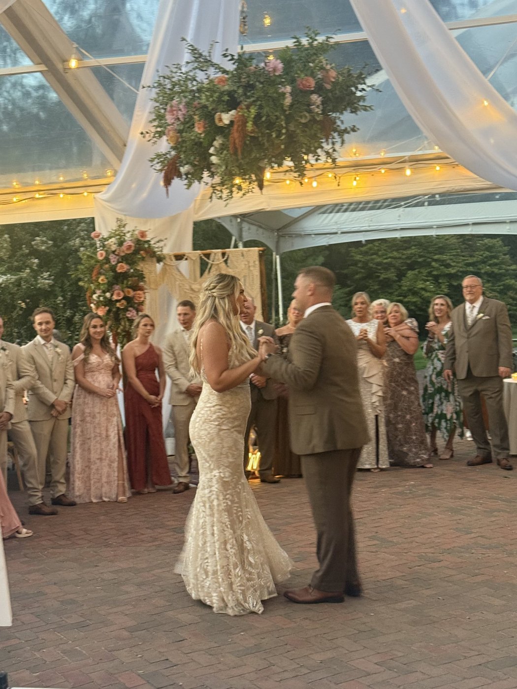 Bride and groom's first dance under hanging florals in a clear tent, guests watching