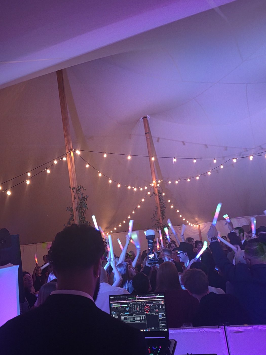 Crowd waving glowing LED sticks under string lights in a tent, seen over Barry's shoulder at the DJ booth