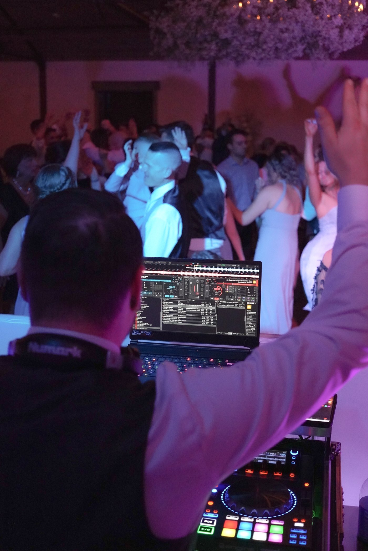 Over Barry's shoulder at the decks — laptop and controller glowing, wedding crowd with hands up